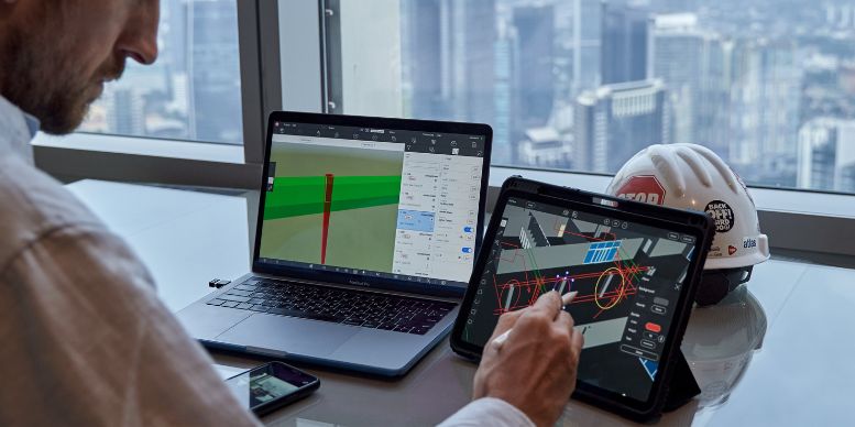  A person working with BIM (Building Information Modeling) software on a laptop and a tablet in an office with a city view. The laptop screen displays a 3D model with structural details, while the tablet shows a more detailed view of the building's components. A smartphone and a construction helmet with stickers are also on the desk.